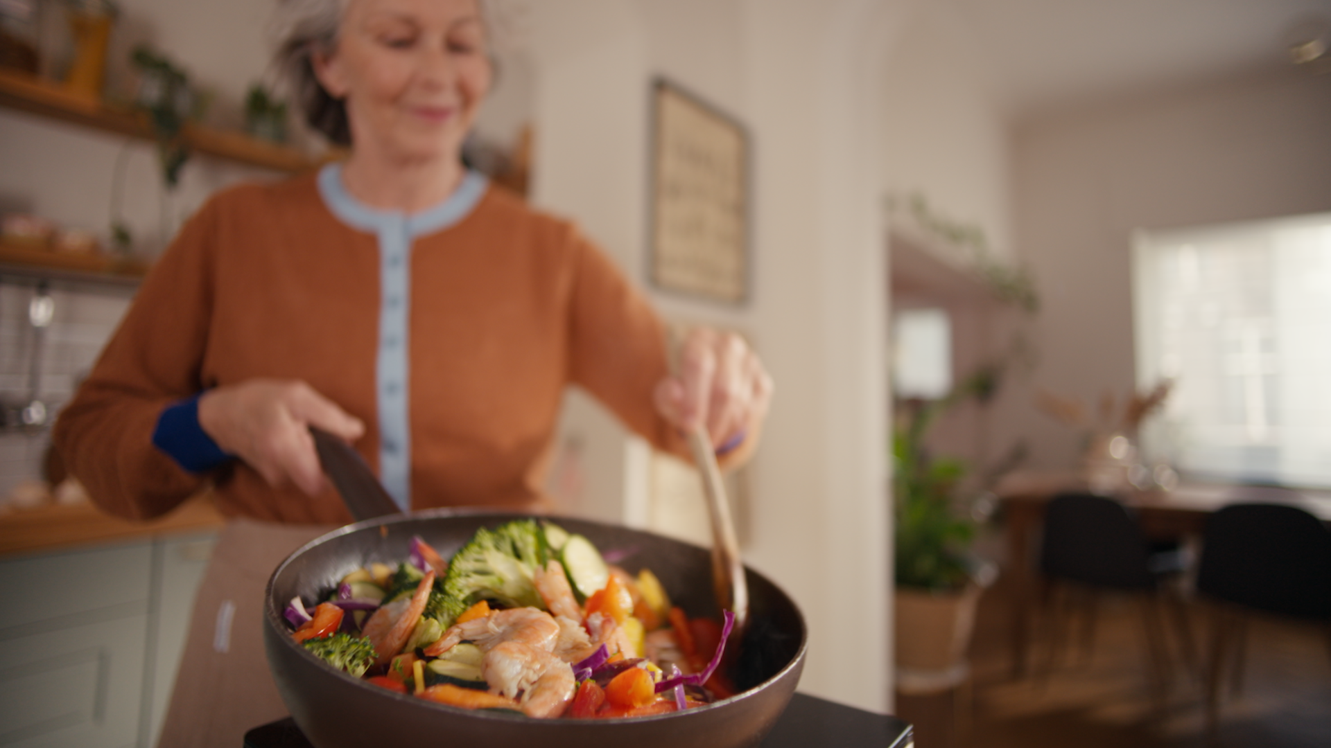 Усмихната жена приготвя скариди в тиган, докато в кухнята се разнасят аромати от готвене