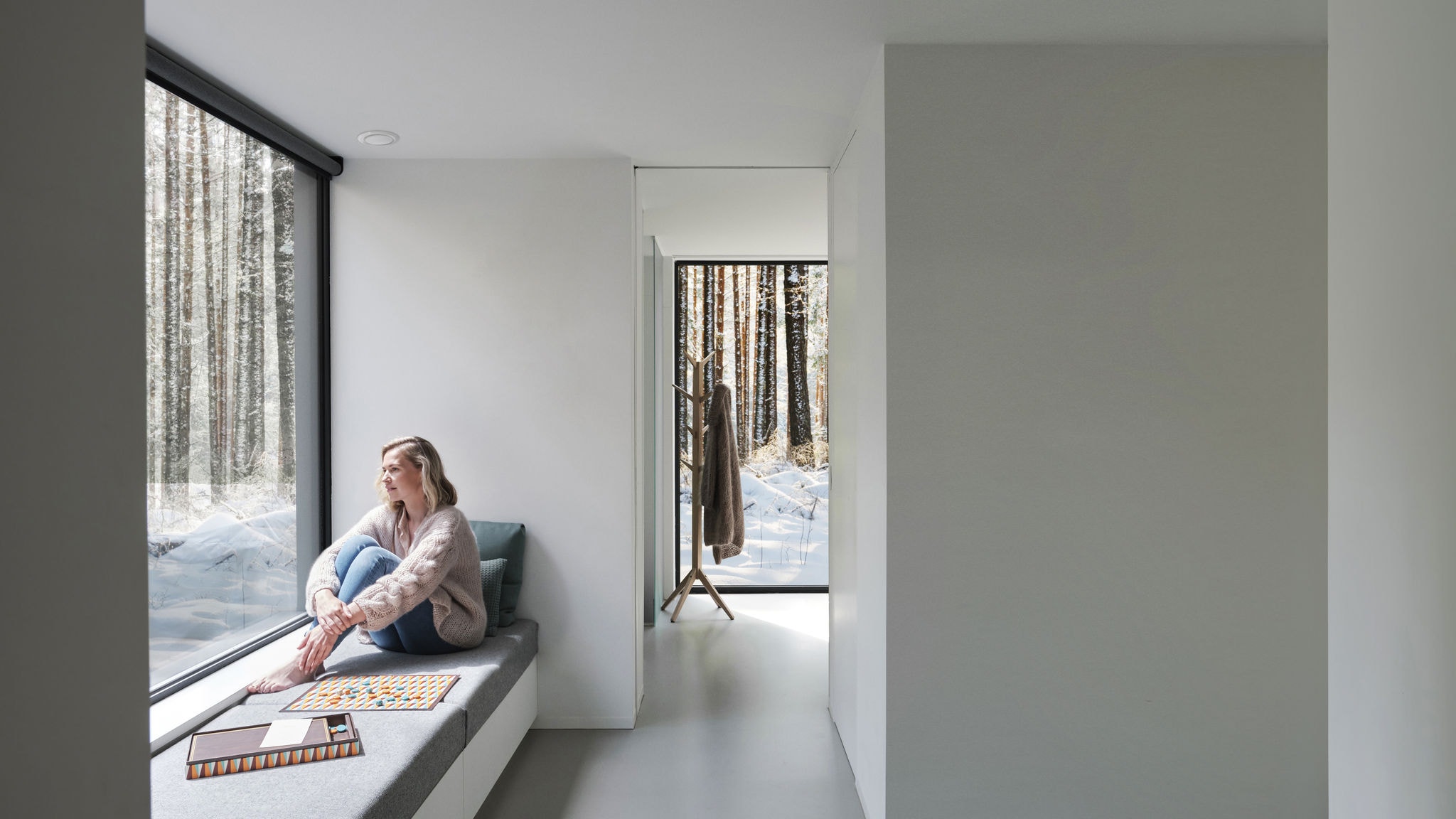 Широкоъгълна снимка на жена, седяща пред прозореца боса, гледаща към дърветата и снега.