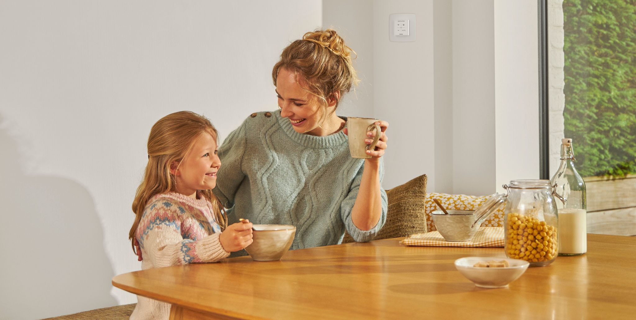 Младо момиче, което яде от купа, седящо на маса до жена с чаша в ръка, и двете усмихнати, щастливи и спокойни, наслаждаващи се на здравословен въздух.